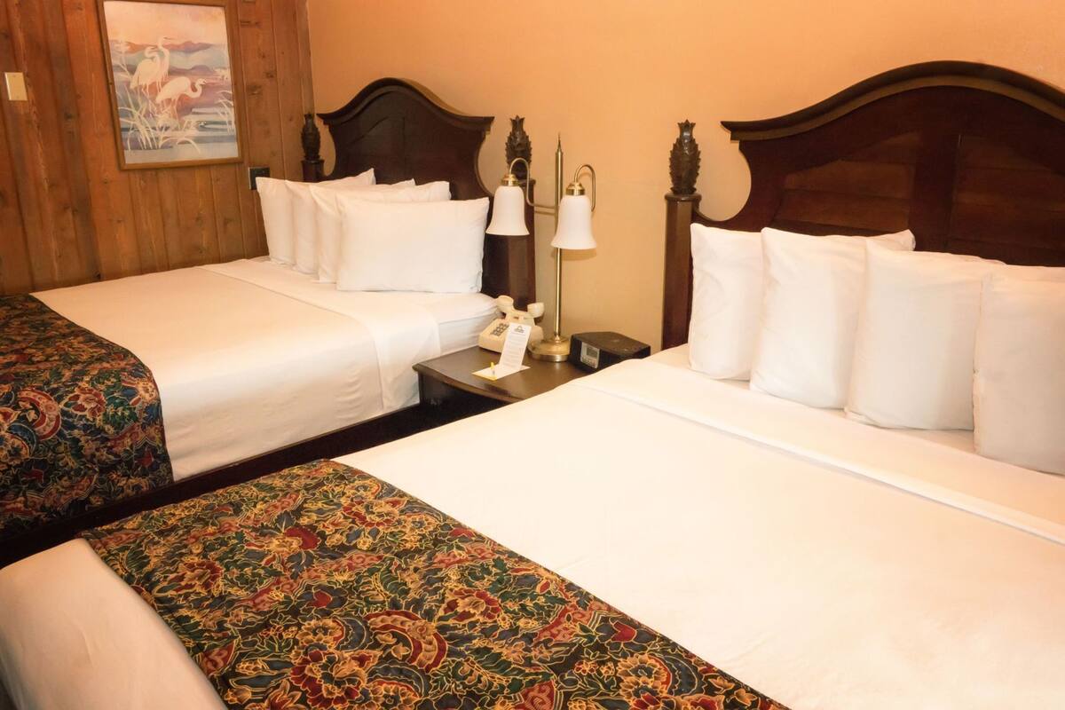 Two double beds with white linens, a wooden headboard, a nightstand with a lamp, and a painting on the wall.