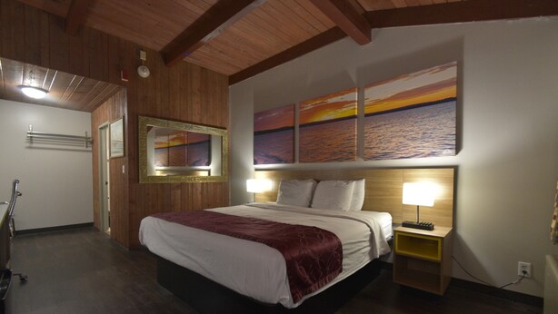 A hotel room with a large bed, wooden ceiling beams, a nightstand with a lamp, and a wall with three sunset paintings. - Days Inn by Wyndham West Allis/Milwaukee (Milwaukee)