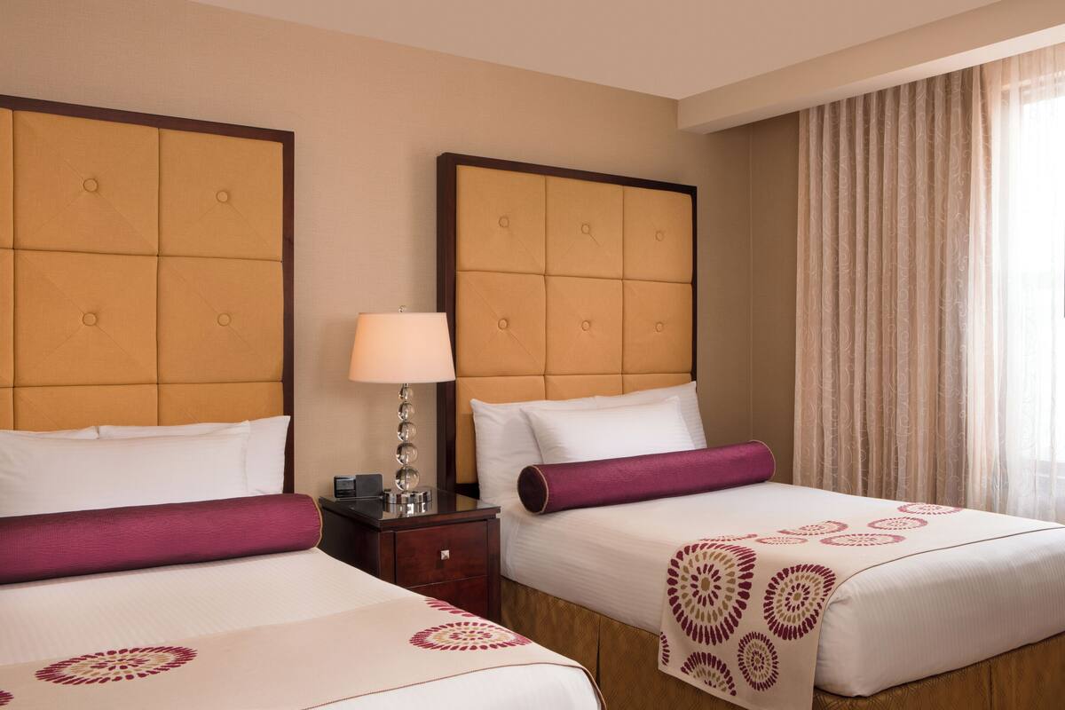 A hotel room with two beds, each with a headboard, a bedside table with a lamp, and a window with curtains.
