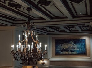 A chandelier with multiple candle-like lights, a ceiling with geometric patterns, and a framed painting of a building by a body of water.