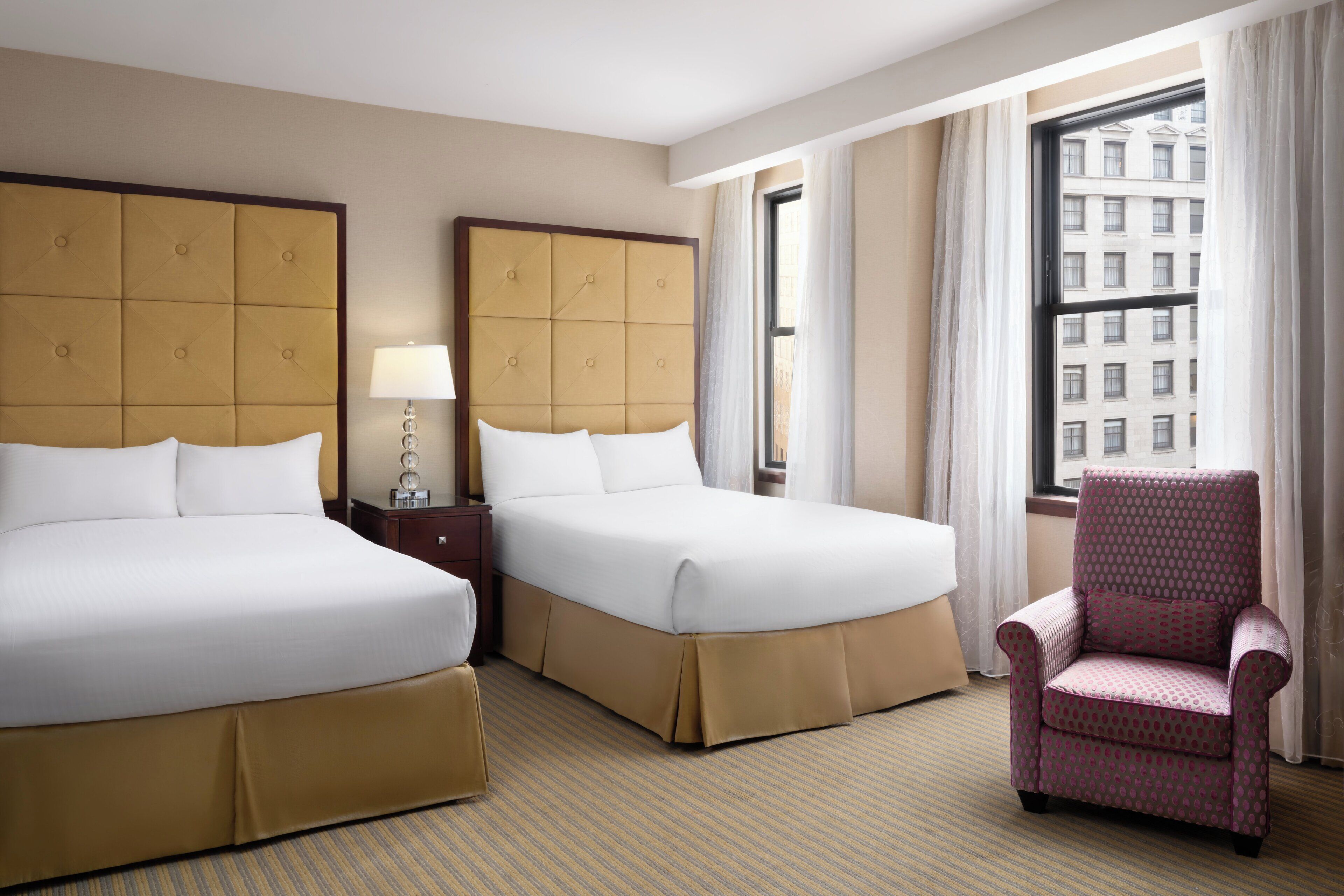 A hotel room with two beds, a chair, a lamp, and a window with a city view.