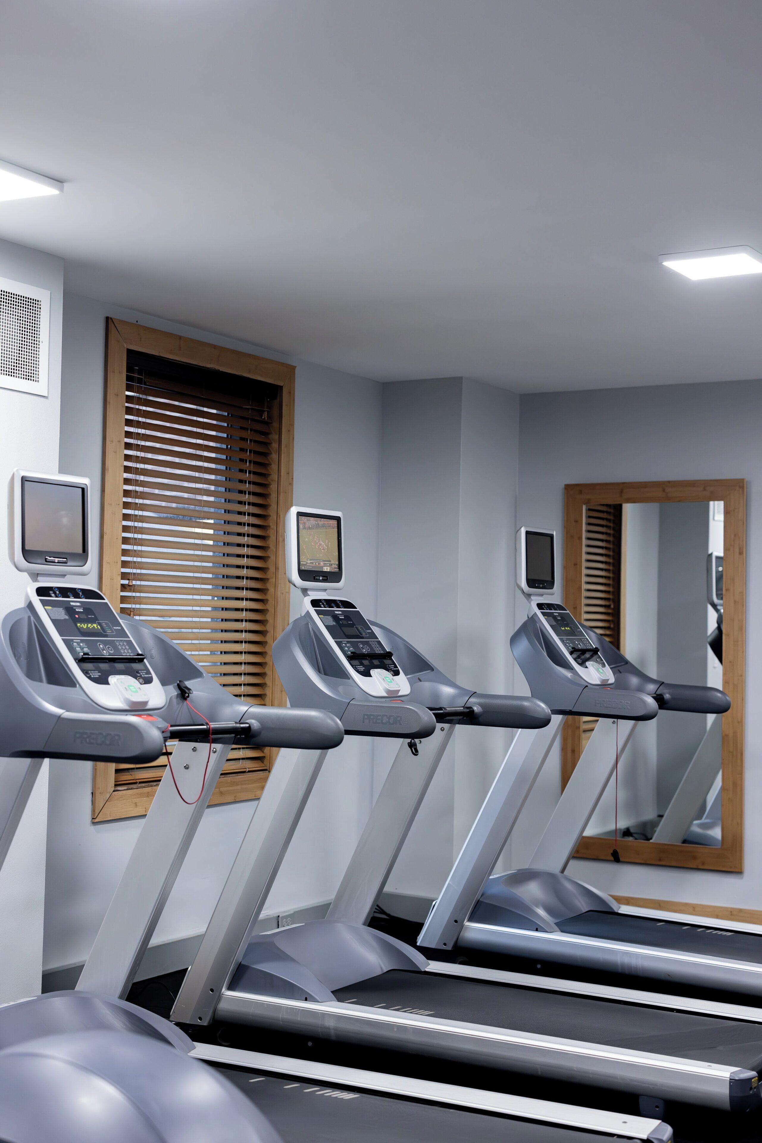 Three treadmills in a gym with wooden blinds and a mirror on the wall.
