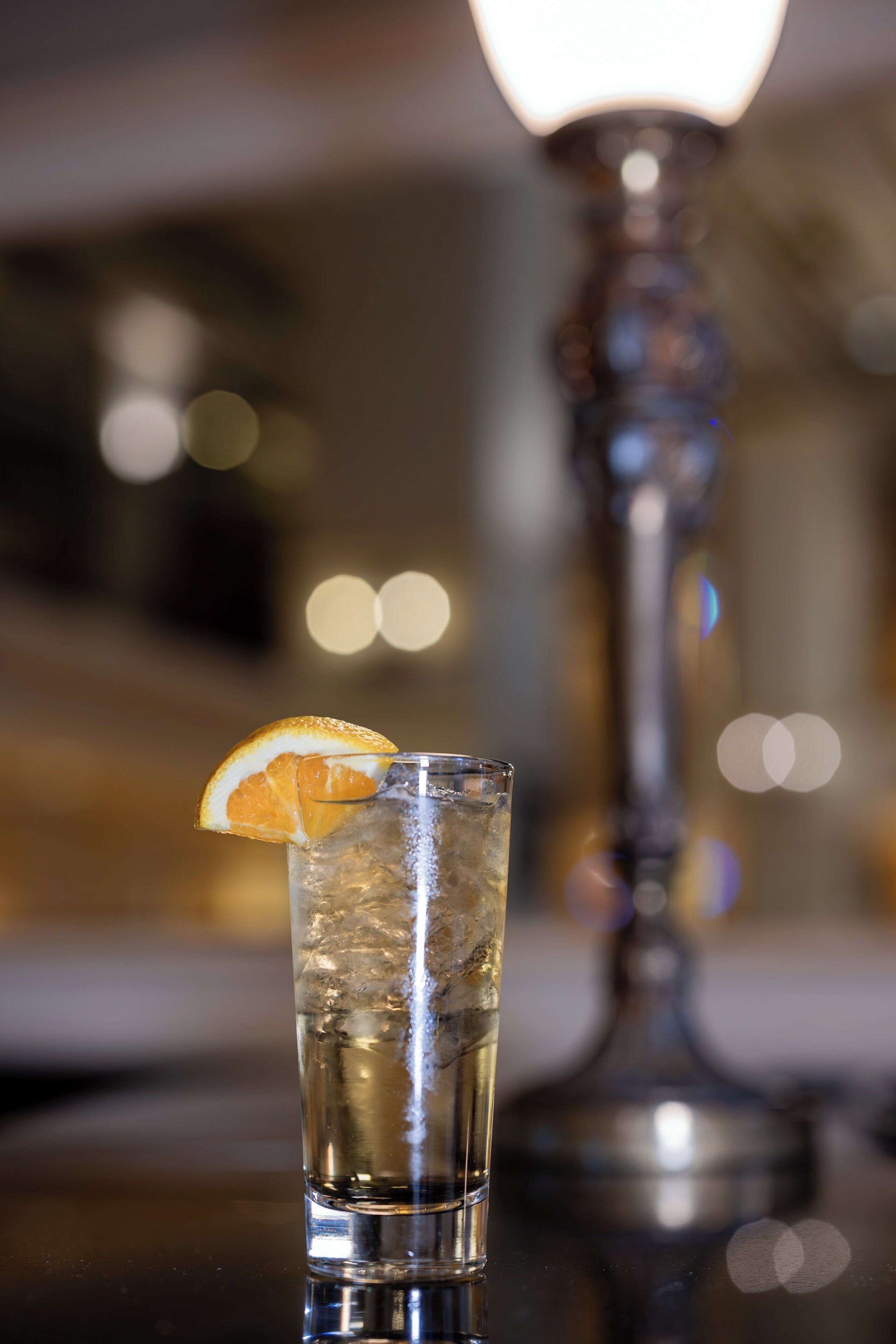 A clear glass with ice and a slice of orange, placed on a reflective surface with a blurred background featuring a tall, ornate lamp.