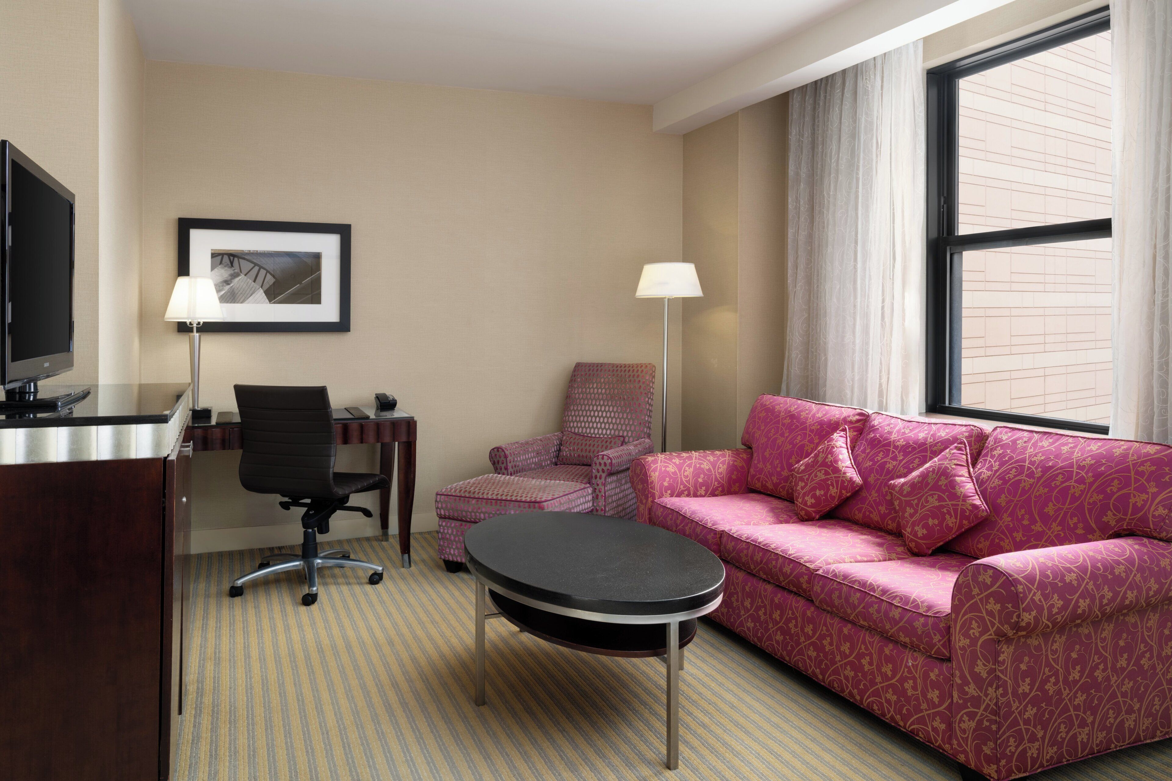 Una habitación de hotel con un escritorio, televisor, sofá estampado rosa, una silla y una lámpara.