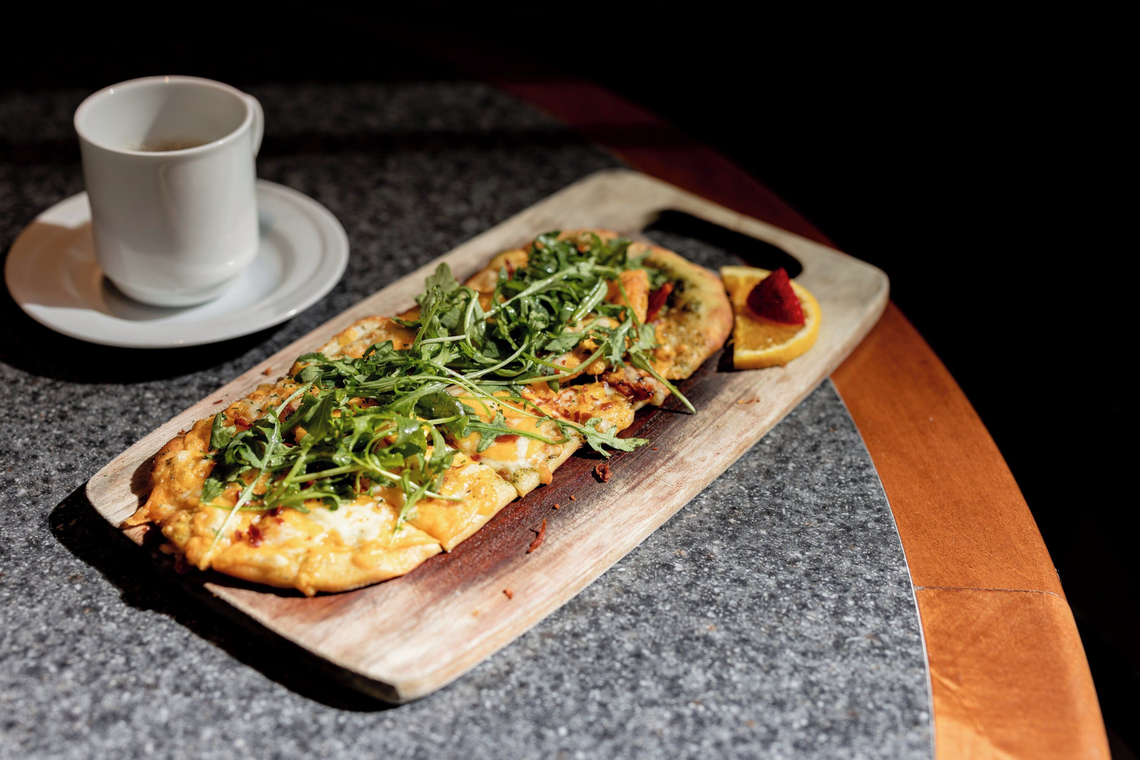 A long, narrow dish with greens and a slice of orange on a wooden board, accompanied by a white cup on a saucer.