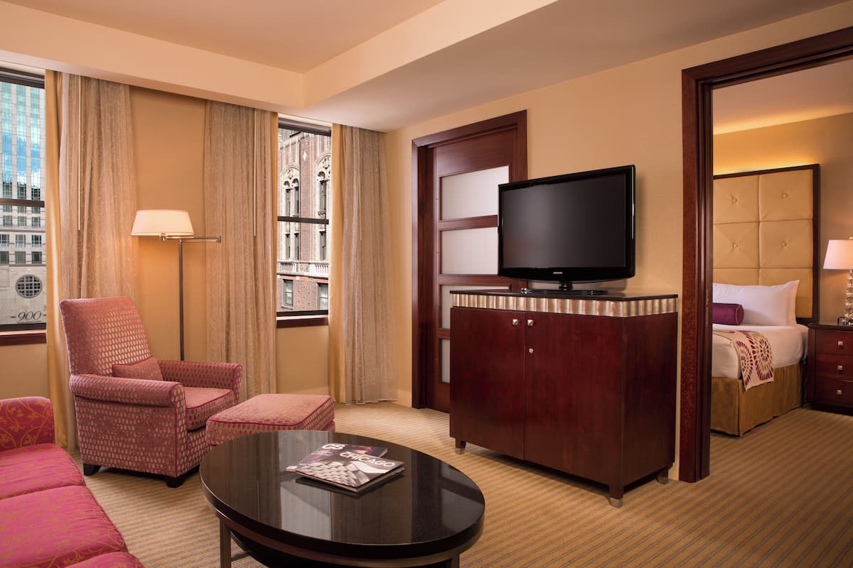 A hotel room with a large window, a television on a wooden cabinet, a red armchair, a sofa, and a small table with a magazine.
