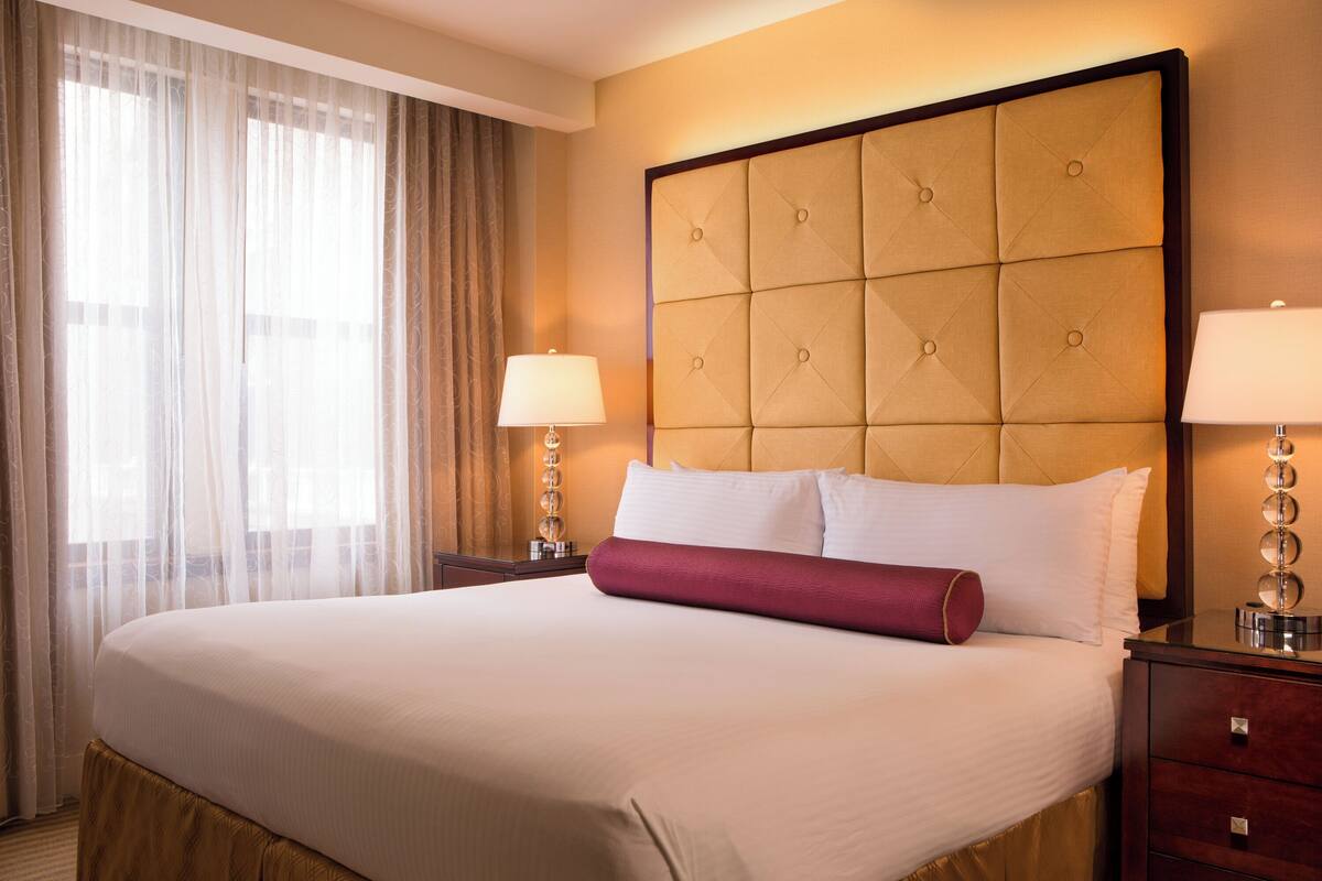 A hotel room with a large bed, two bedside lamps, a wooden nightstand, and a large window with sheer curtains.