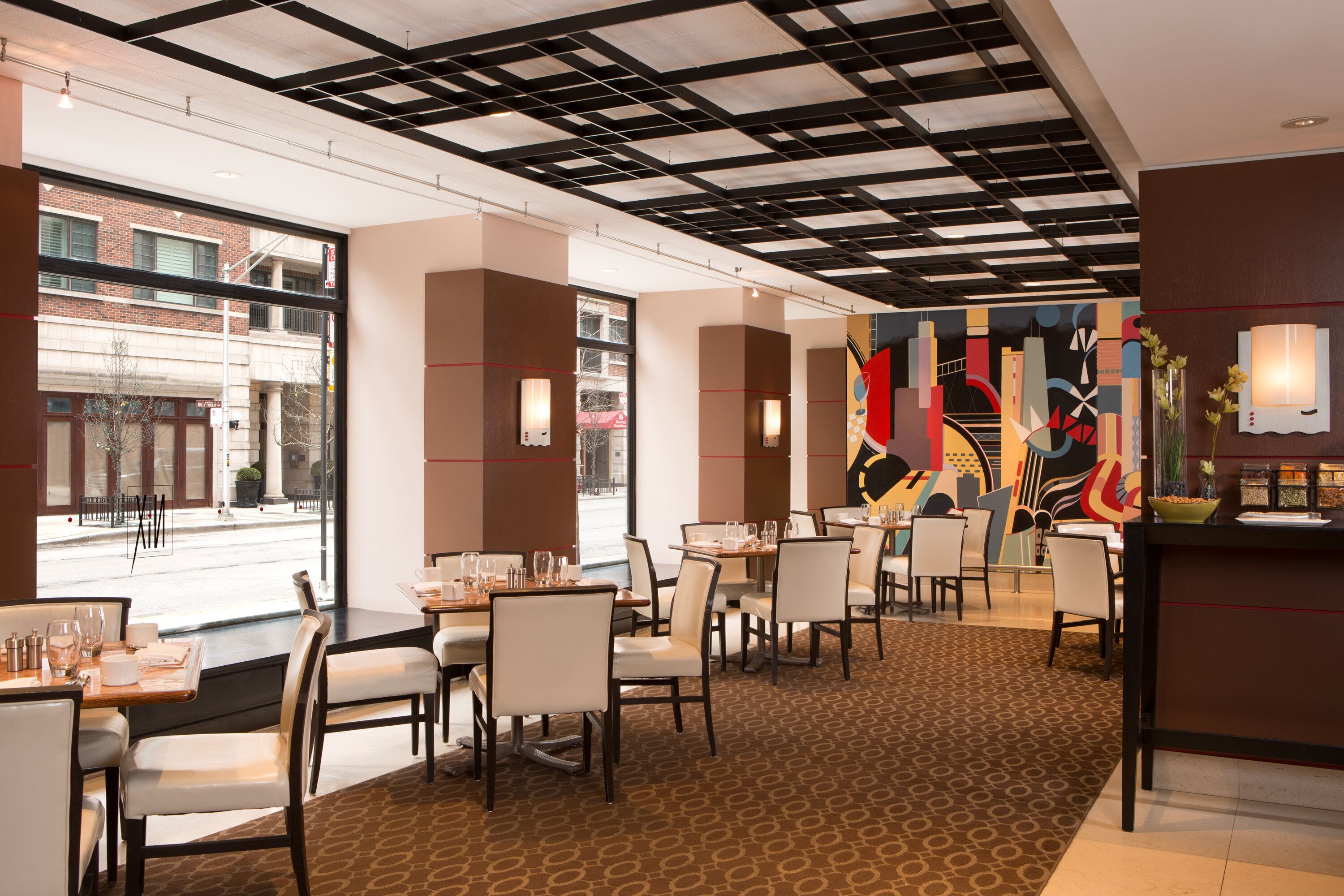 Um restaurante com mesas e cadeiras, um tapete estampado e vista da rua através de grandes janelas.