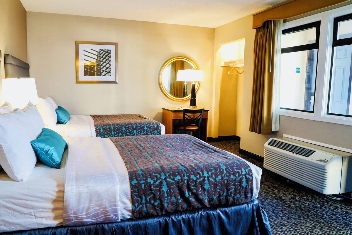 A hotel room with two beds, a desk, a chair, a mirror, a window with curtains, and a wall-mounted air conditioner.
