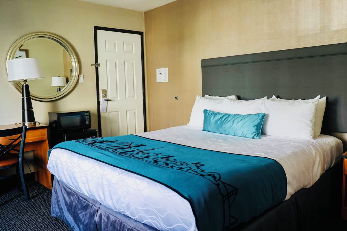 A hotel room with a bed, a desk, a chair, a lamp, a mirror, and a door.