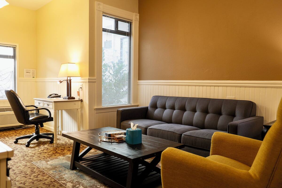 A living room with a dark brown sofa, a yellow armchair, a wooden coffee table, a lamp, and a window with blinds.