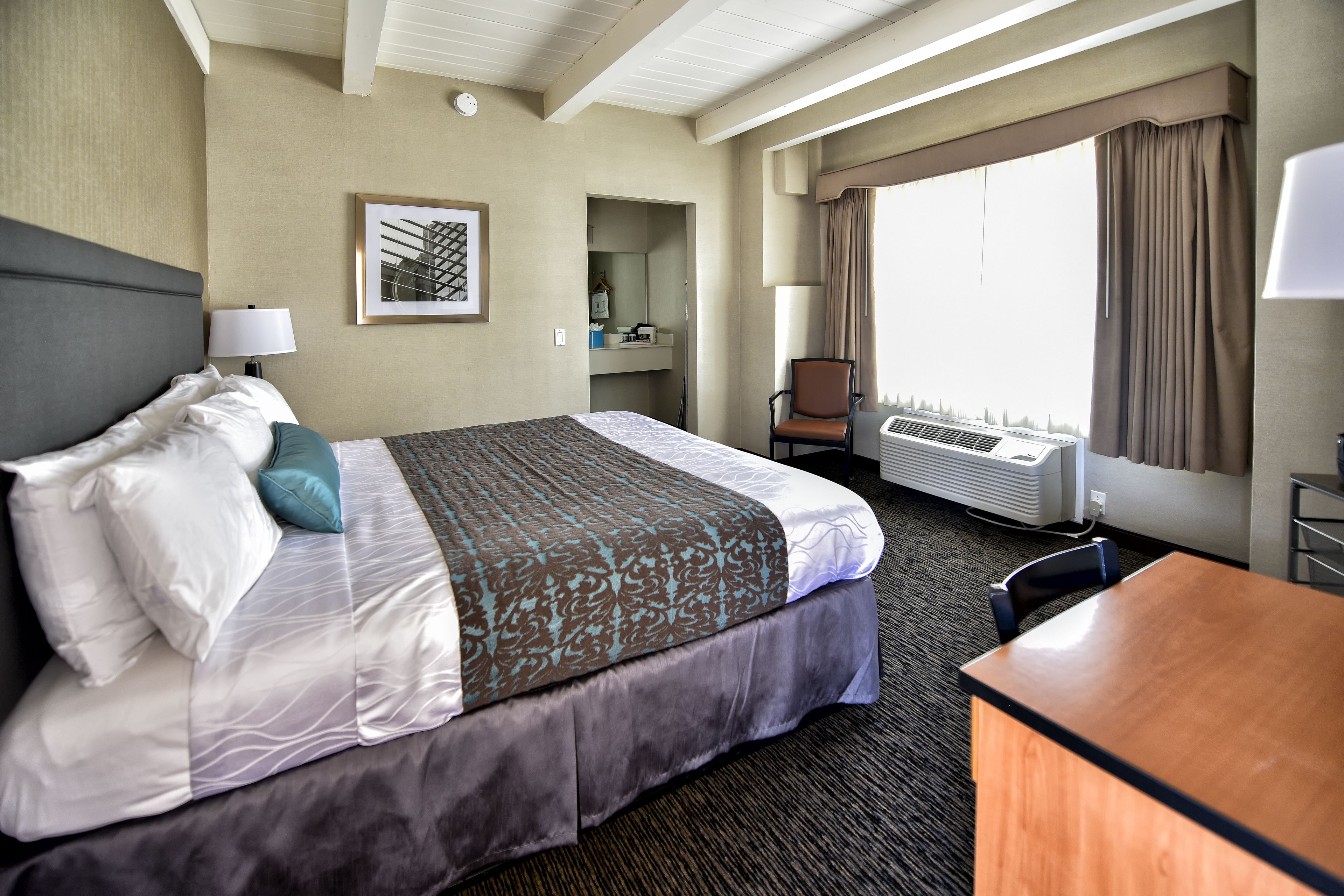 A hotel room with a large bed, a desk, a chair, a window with curtains, a wall-mounted air conditioner, and a framed picture on the wall.