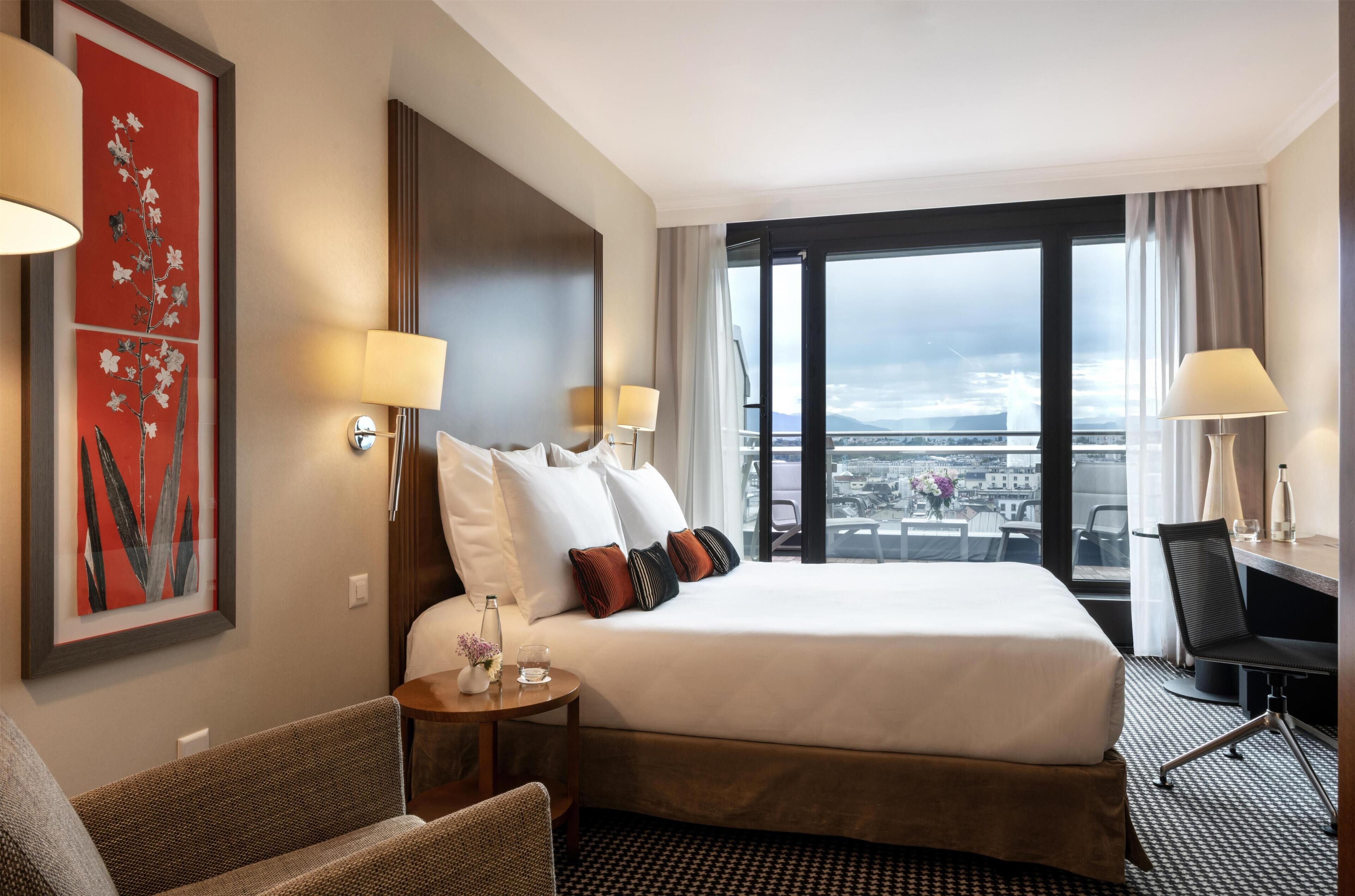 A hotel room with a large bed, two bedside lamps, a small table with a vase and glasses, a chair, and a view of a cityscape through a large window.
