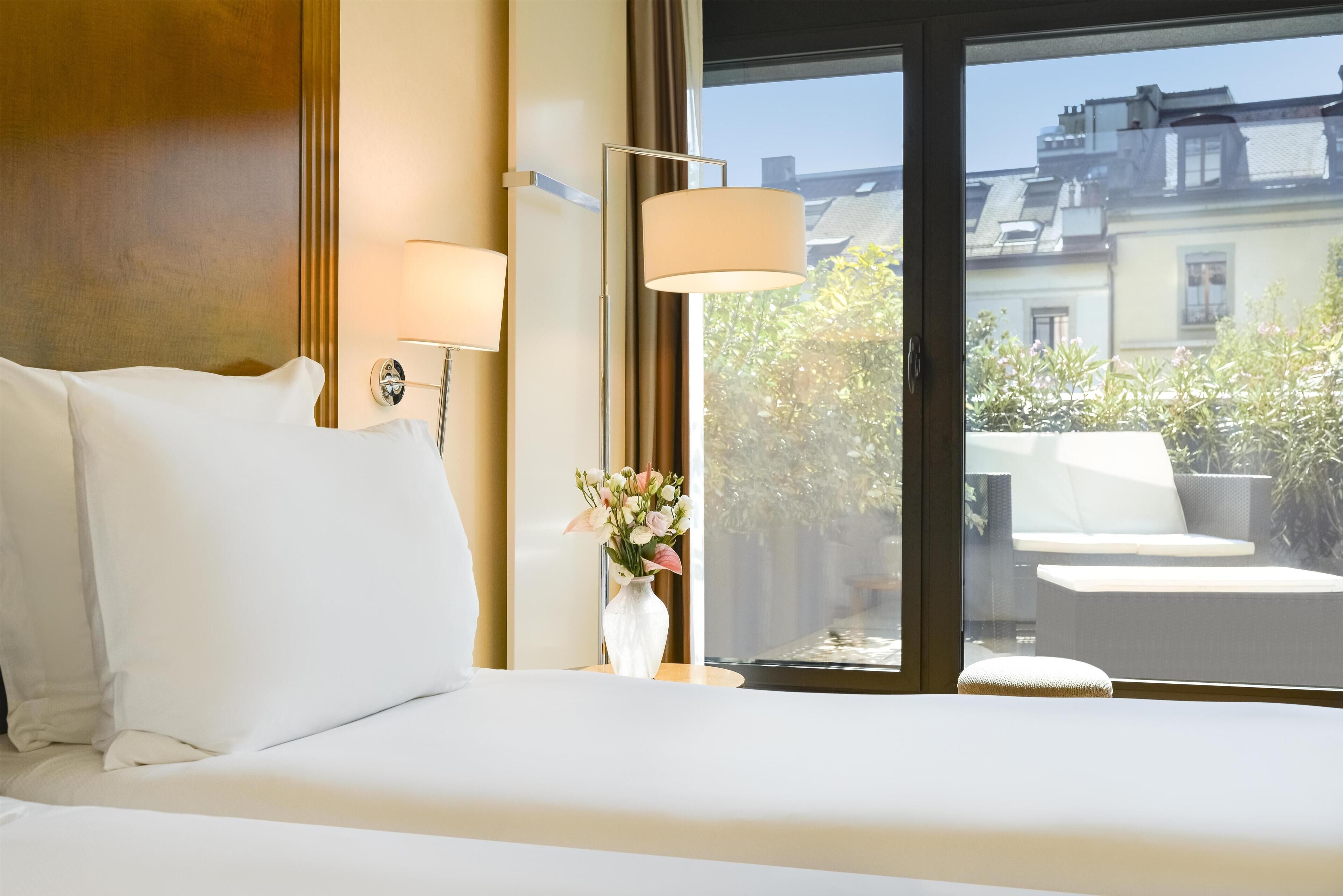 A hotel room with a bed, two wall-mounted lamps, a vase with flowers, and a view of the outside.