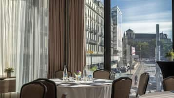 A hotel room with a dining table set for two, a view of a city street, and a large window with curtains.
