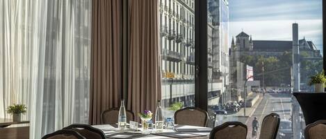 A hotel room with a dining table set for two, a view of a city street, and a large window with curtains.