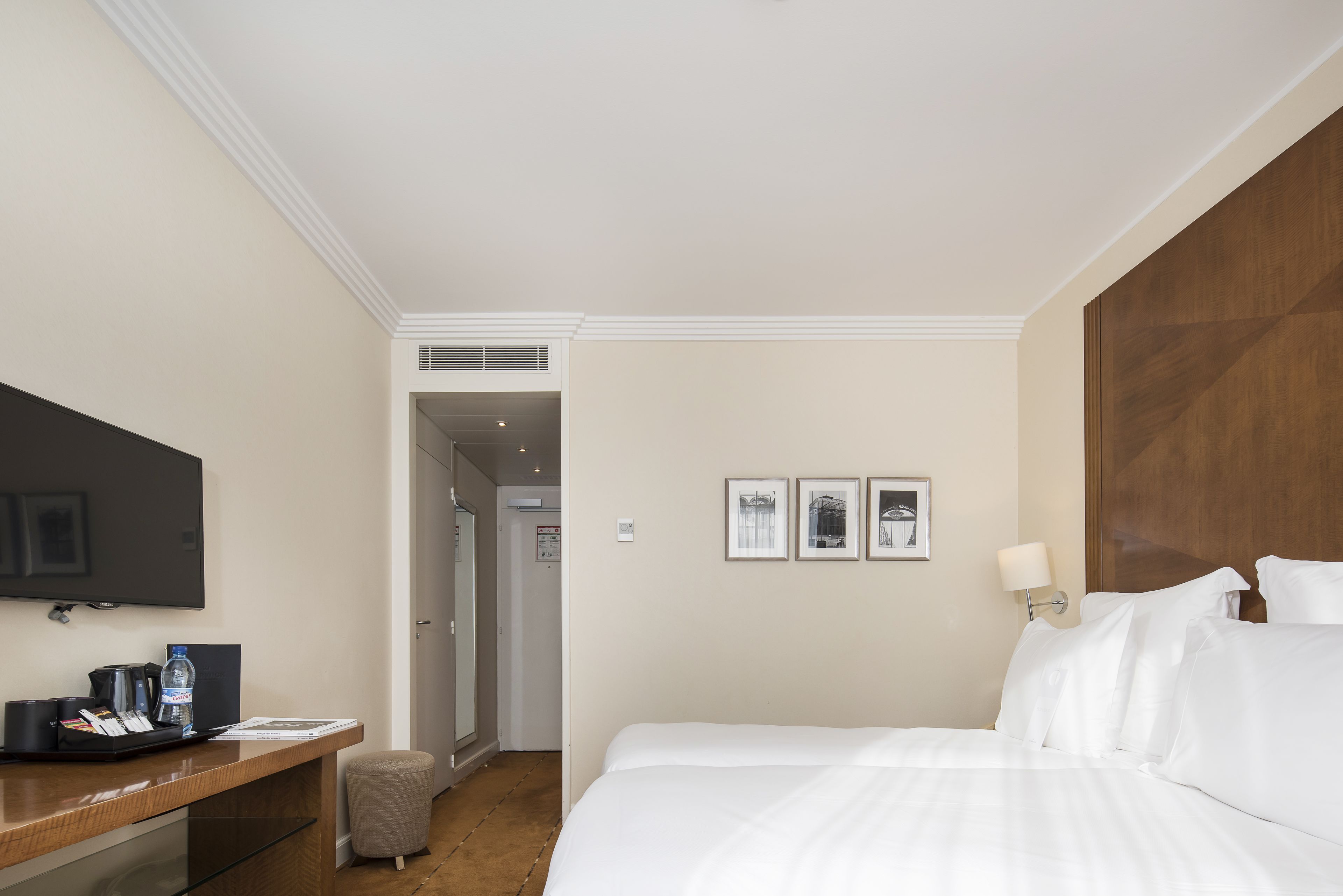 A hotel room with a bed, a desk with a flat-screen TV, a coffee maker, and a water bottle.