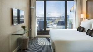 A hotel room with a large bed, a glass desk, a flat-screen TV, and a city view through the windows. - Warwick Geneva (Geneva)