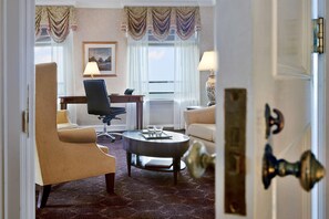 A hotel room with a beige armchair, a dark-colored sofa, a round coffee table with a book and glasses, a desk with a lamp, and a view through the window.