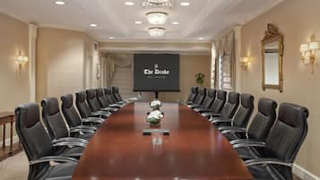 A conference room with a long table, black leather chairs, chandeliers, and a screen displaying "The Drake Real Chicago".
