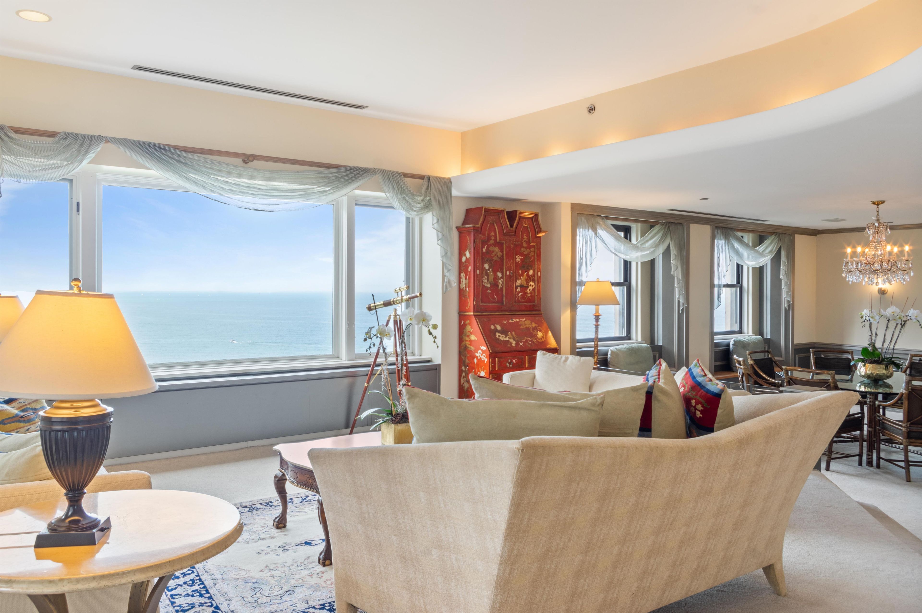 A hotel room with a large window offering a view of the sea, a sitting area with a sofa and armchairs, a dining table with chairs, and a chandelier.