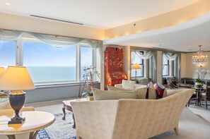 A hotel room with a large window offering a view of the sea, a sitting area with a sofa and armchairs, a dining table with chairs, and a chandelier.