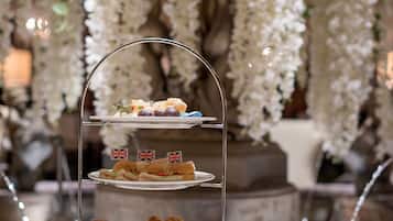 A three-tiered dessert stand with various pastries and sandwiches, surrounded by tea sets and a teapot on a round table, set against a backdrop of hanging white flowers and a stone fountain.