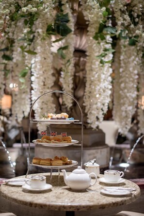A three-tiered dessert stand with various pastries and sandwiches, surrounded by tea sets and a teapot on a round table, set against a backdrop of hanging white flowers and a stone fountain.