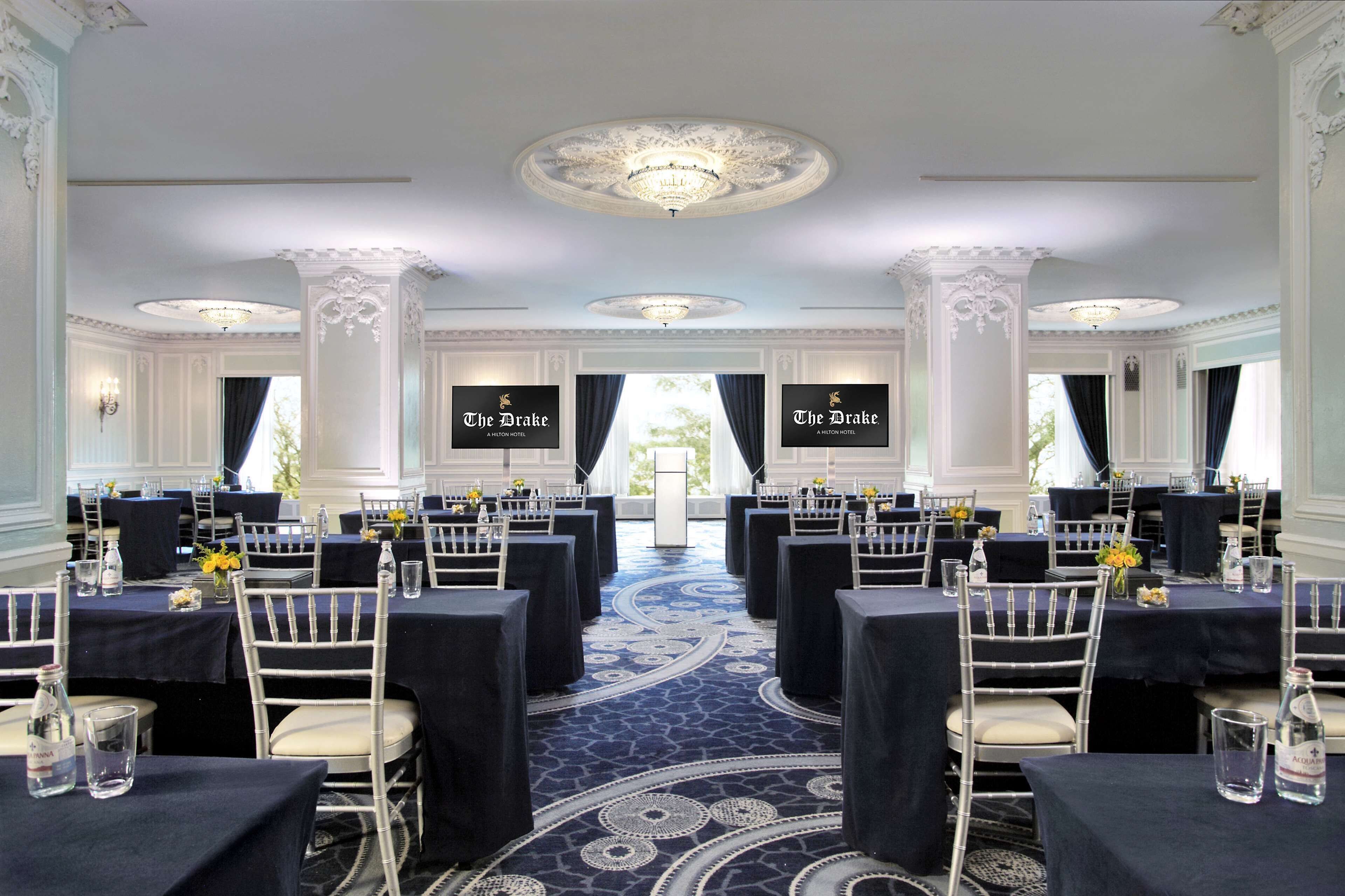 A formal dining room with tables set for a banquet, featuring white chairs and dark tablecloths.