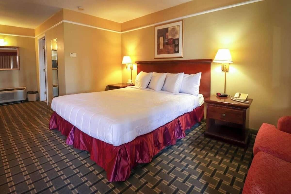 Quarto de hotel com uma cama grande, duas mesas de cabeceira, um criado-mudo, uma cadeira vermelha e uma porta que dá para outro quarto.