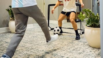 To personer spiller fodbold indendørs med et mål, en lysekrone og potteplanter.