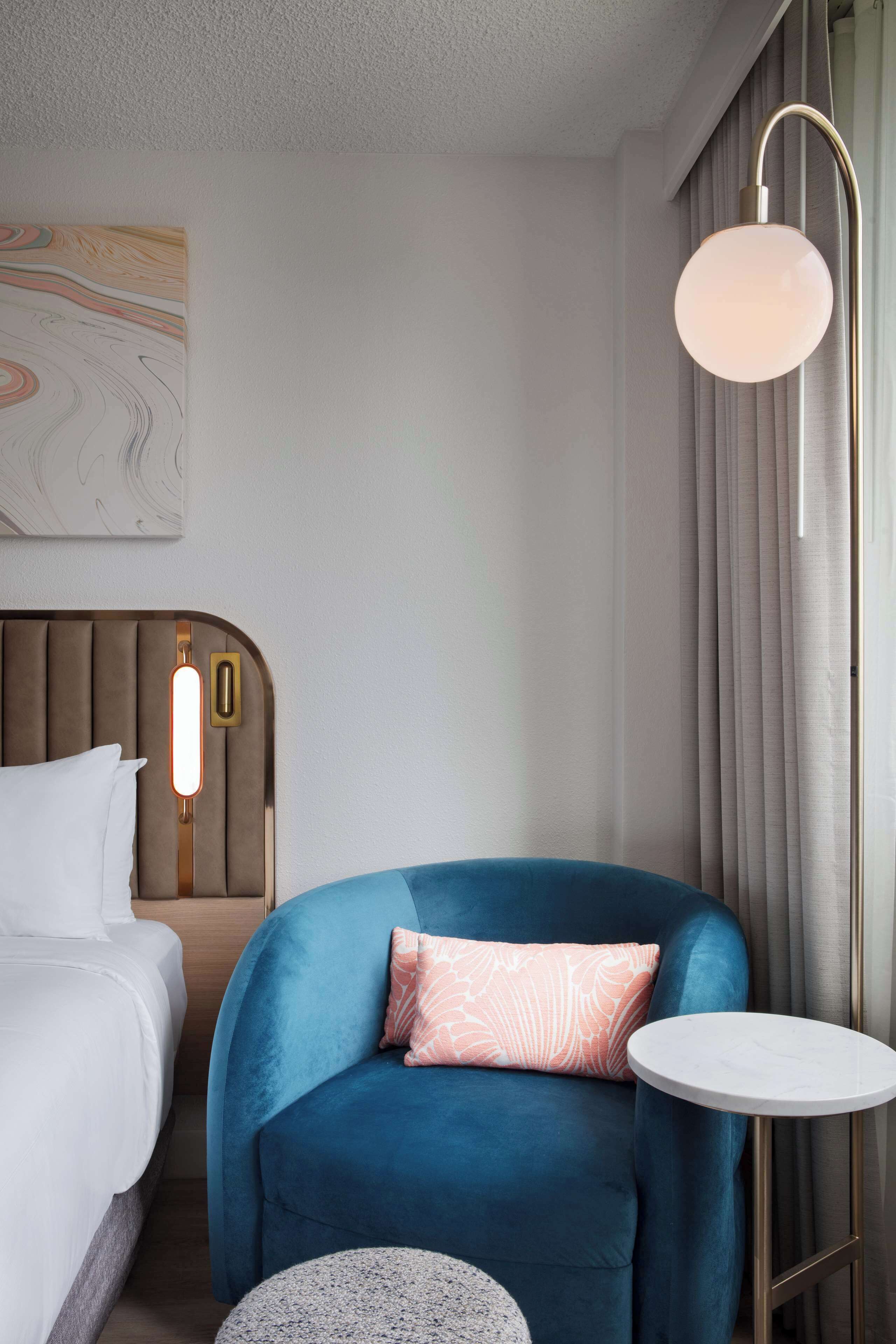 Una habitación de hotel con una cama, un sillón azul, una mesita redonda y una lámpara fijada a la pared.
