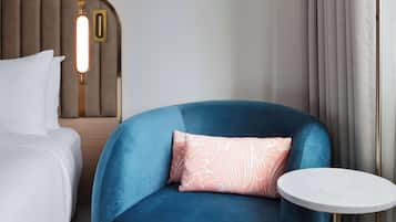 Una habitación de hotel con una cama, un sillón azul, una mesita redonda y una lámpara fijada a la pared.