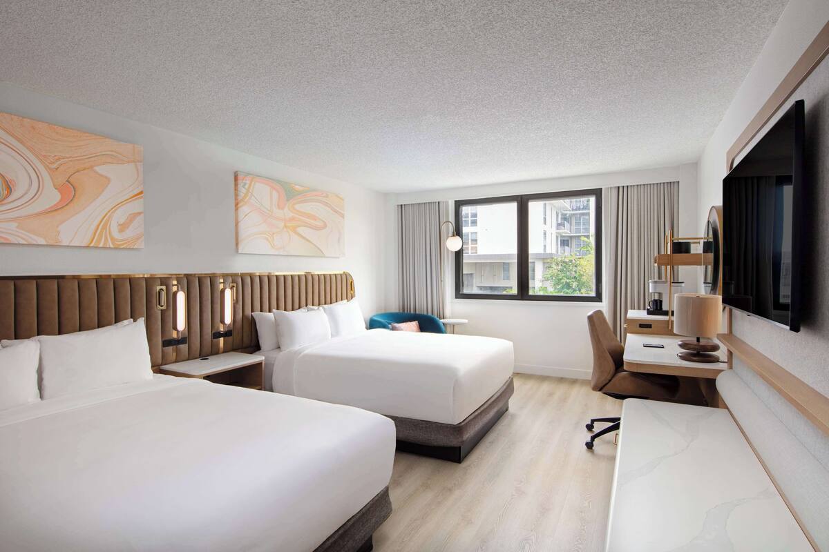 A hotel room with two beds, a desk, a chair, a TV, and artwork on the walls.