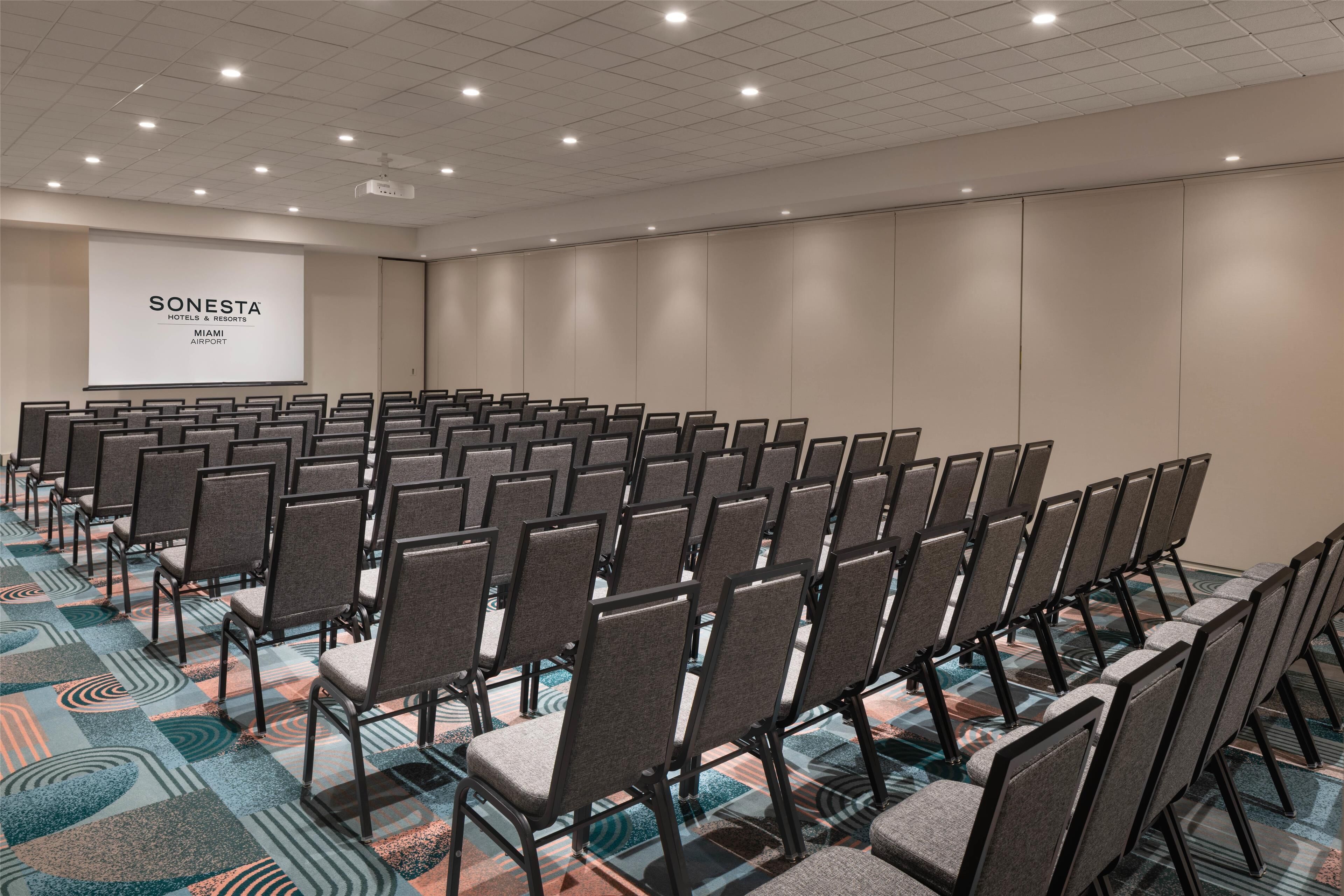 Une salle de conférence avec des rangées de chaises, un écran de projection affichant le texte « SONESTA HOTELS & RESORTS MIAMI AIRPORT », et une moquette à motifs.