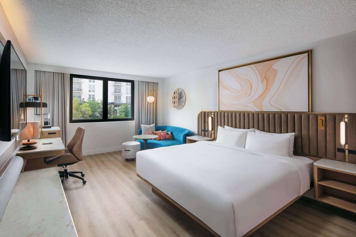 A modern hotel room with a large bed, a desk with a chair, a small round table, a blue sofa, and a large window offering a view of buildings.
