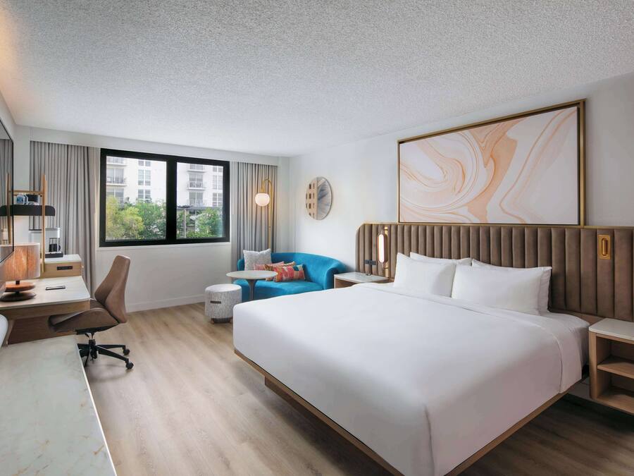 A modern hotel room with a large bed, a desk with a chair, a small round table, a blue sofa, and a large window offering a view of buildings.