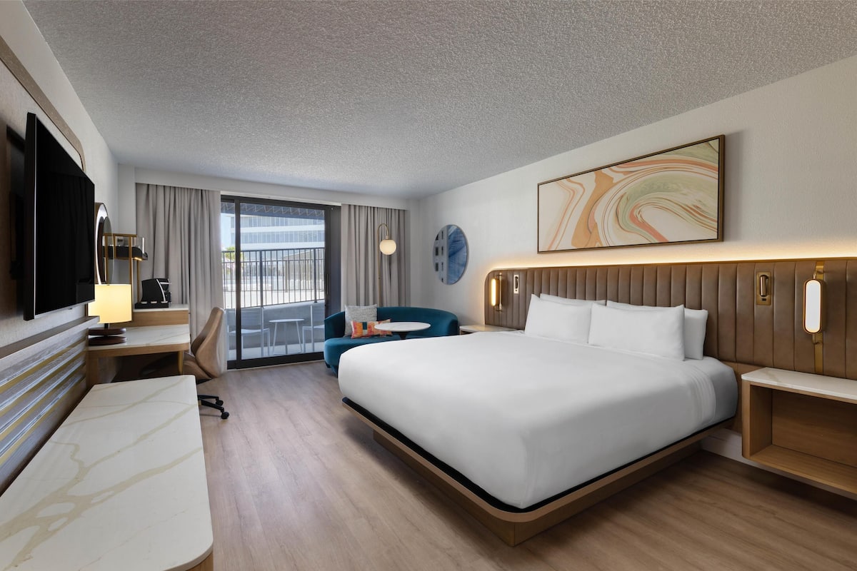 A modern hotel room with a large bed, a desk, a chair, a TV, and a balcony with curtains.