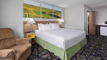 Una habitación de hotel con una cama grande, dos lámparas de noche, un cabecero de madera, un cubrelecho verde y un sillón marrón. En la pared, un cuadro de un campo.