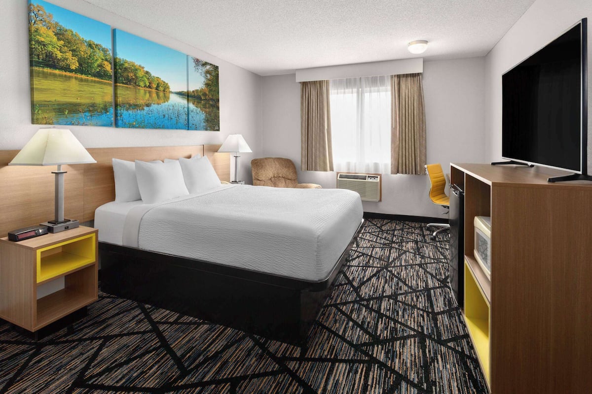 Une chambre d’hôtel avec un grand lit, un bureau doté d’un téléviseur à écran plat, une chaise, une lampe et un tableau représentant un paysage fluvial accroché au mur.
