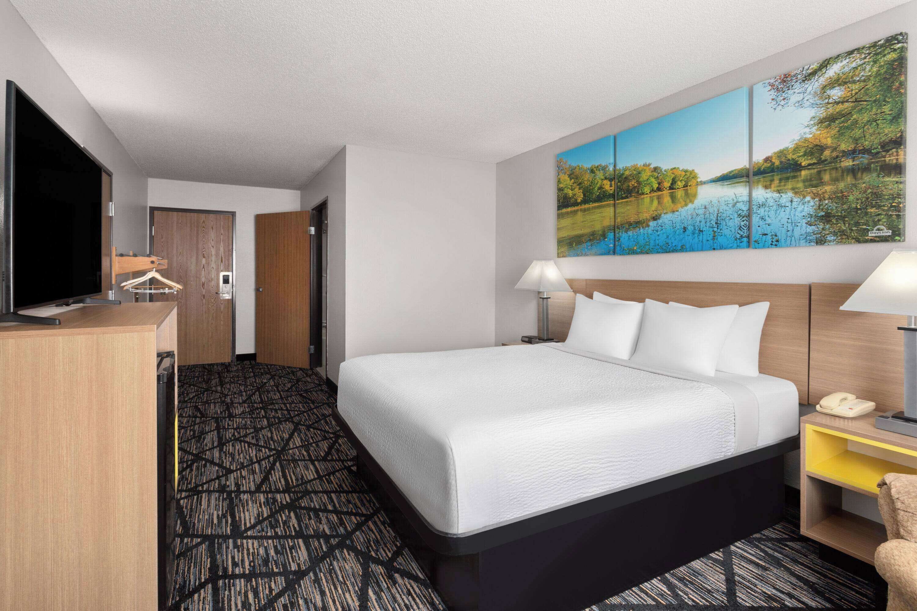 A hotel room with a large bed, a desk with a TV, a painting of a lake, and a lamp.