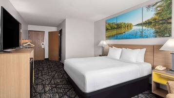 A hotel room with a large bed, a desk with a TV, a painting of a lake, and a lamp.