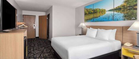 A hotel room with a large bed, a desk with a TV, a painting of a lake, and a lamp.