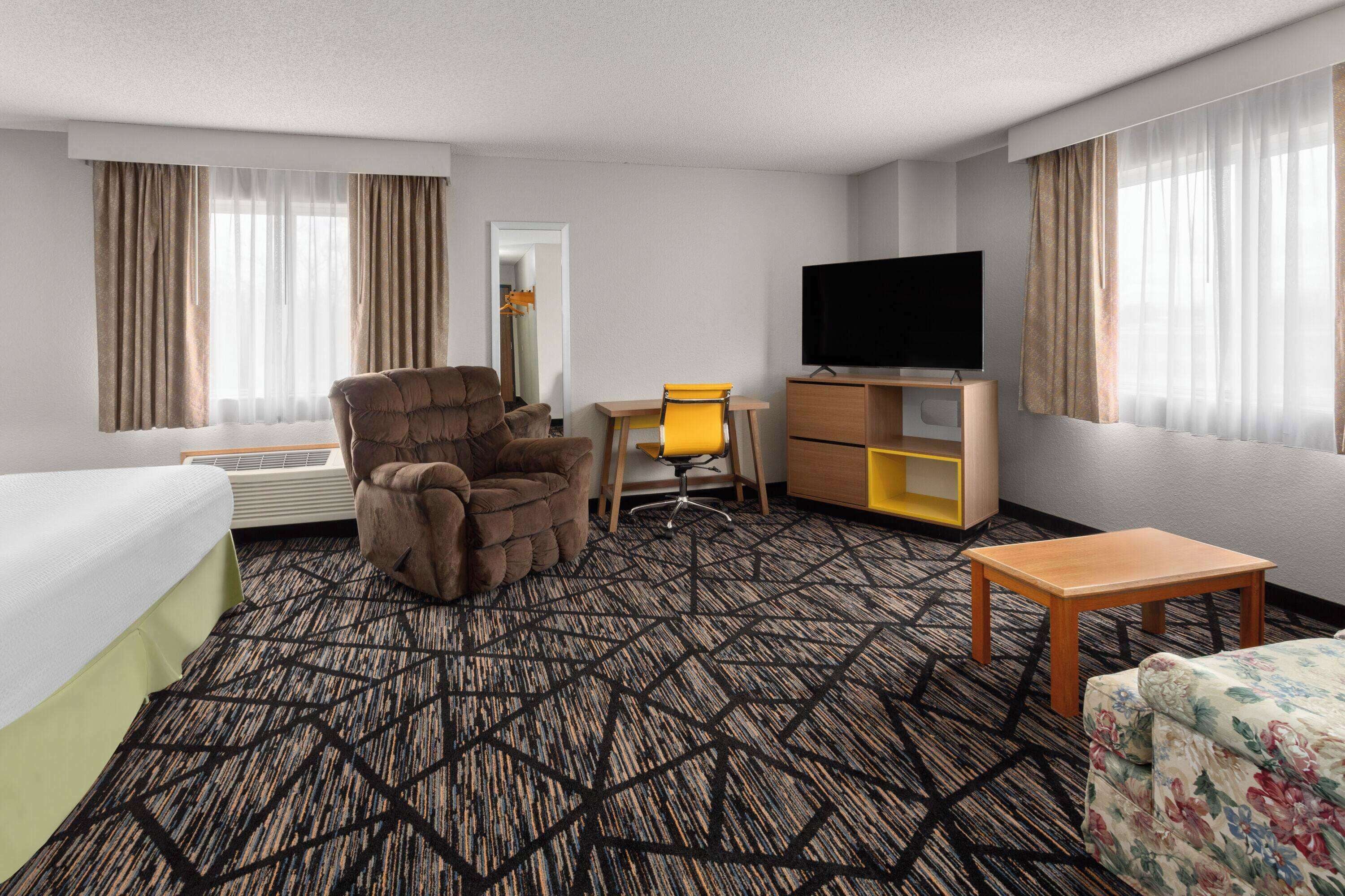 A hotel room with a bed, a brown armchair, a desk with a chair, a television, a small table, and a sofa.