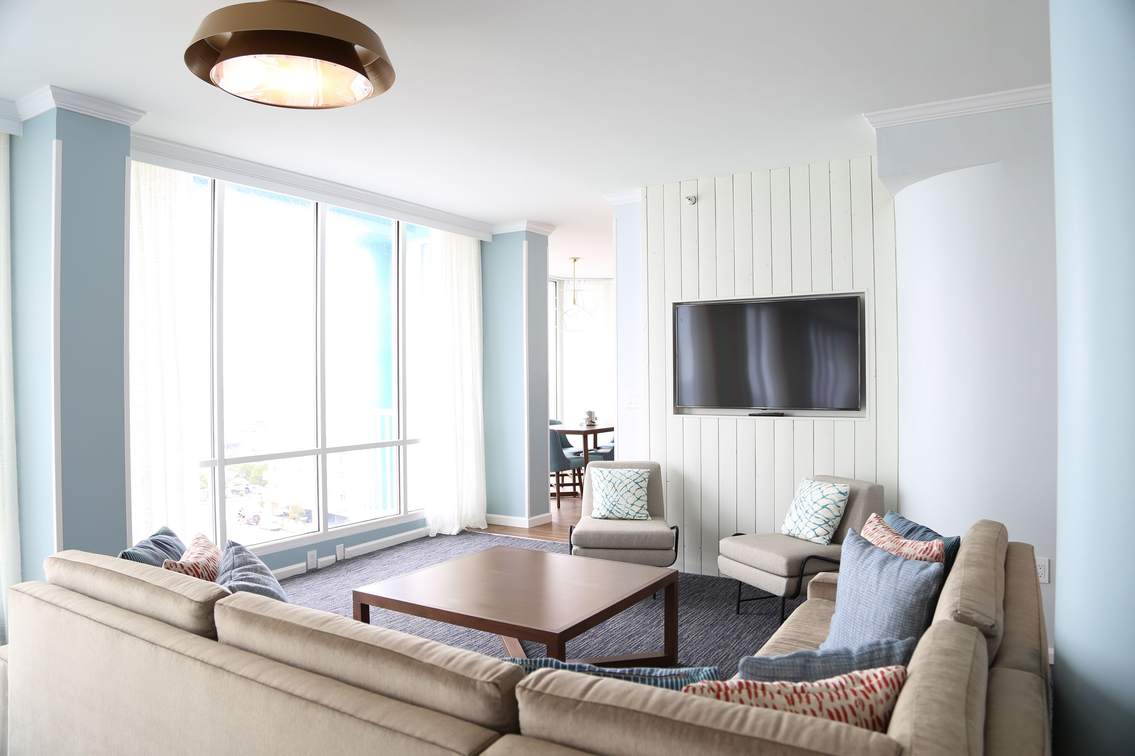 A spacious living room with a large window, a flat-screen TV mounted on the wall, a beige sofa, and a coffee table.