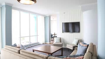 A spacious living room with a large window, a flat-screen TV mounted on the wall, a beige sofa, and a coffee table.