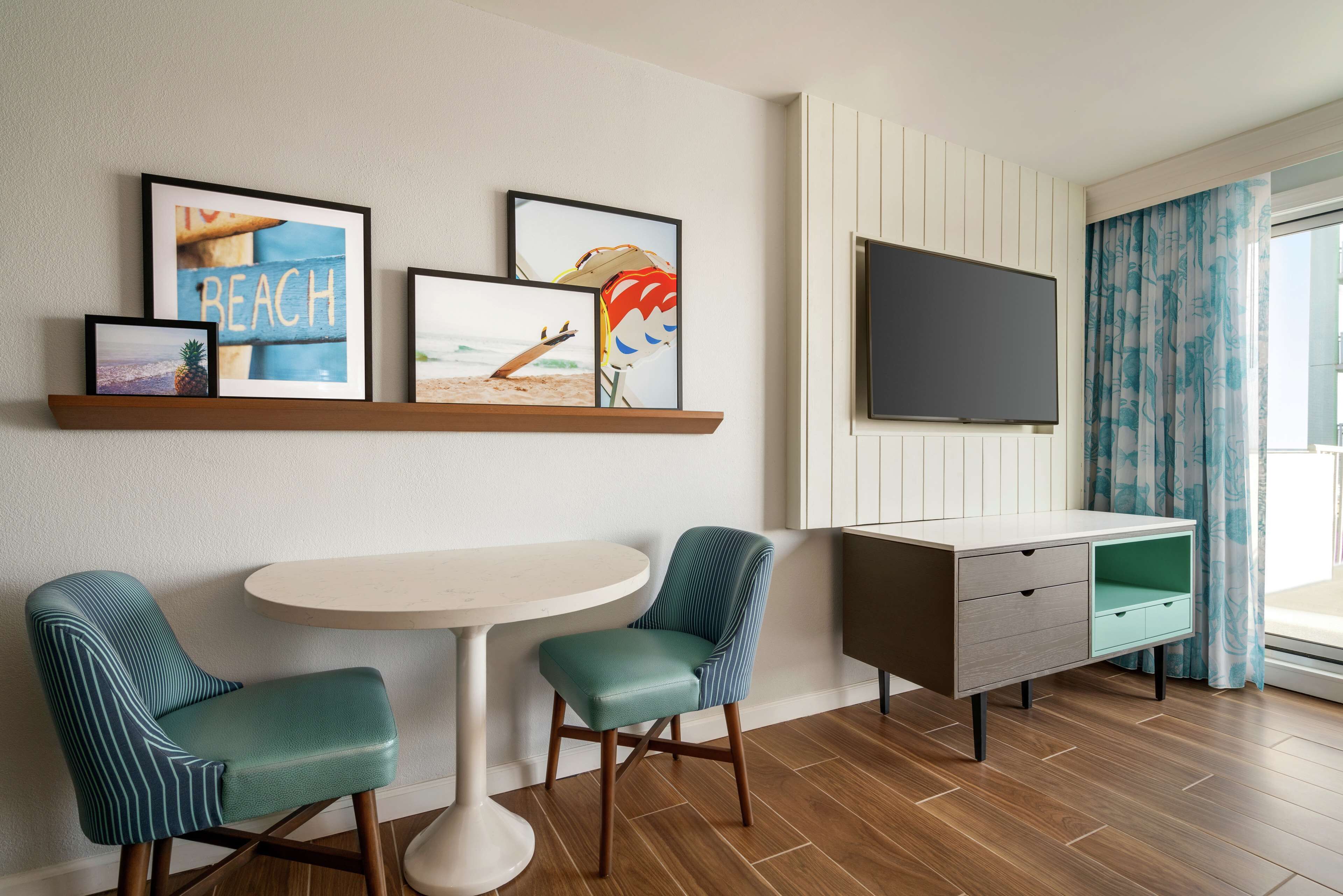 Habitación de hotel con piso de madera, una mesa redonda, dos sillas color turquesa, televisor, mueble y obras de arte con temática de playa.