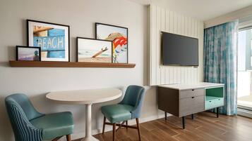 Habitación de hotel con piso de madera, una mesa redonda, dos sillas color turquesa, televisor, mueble y obras de arte con temática de playa.