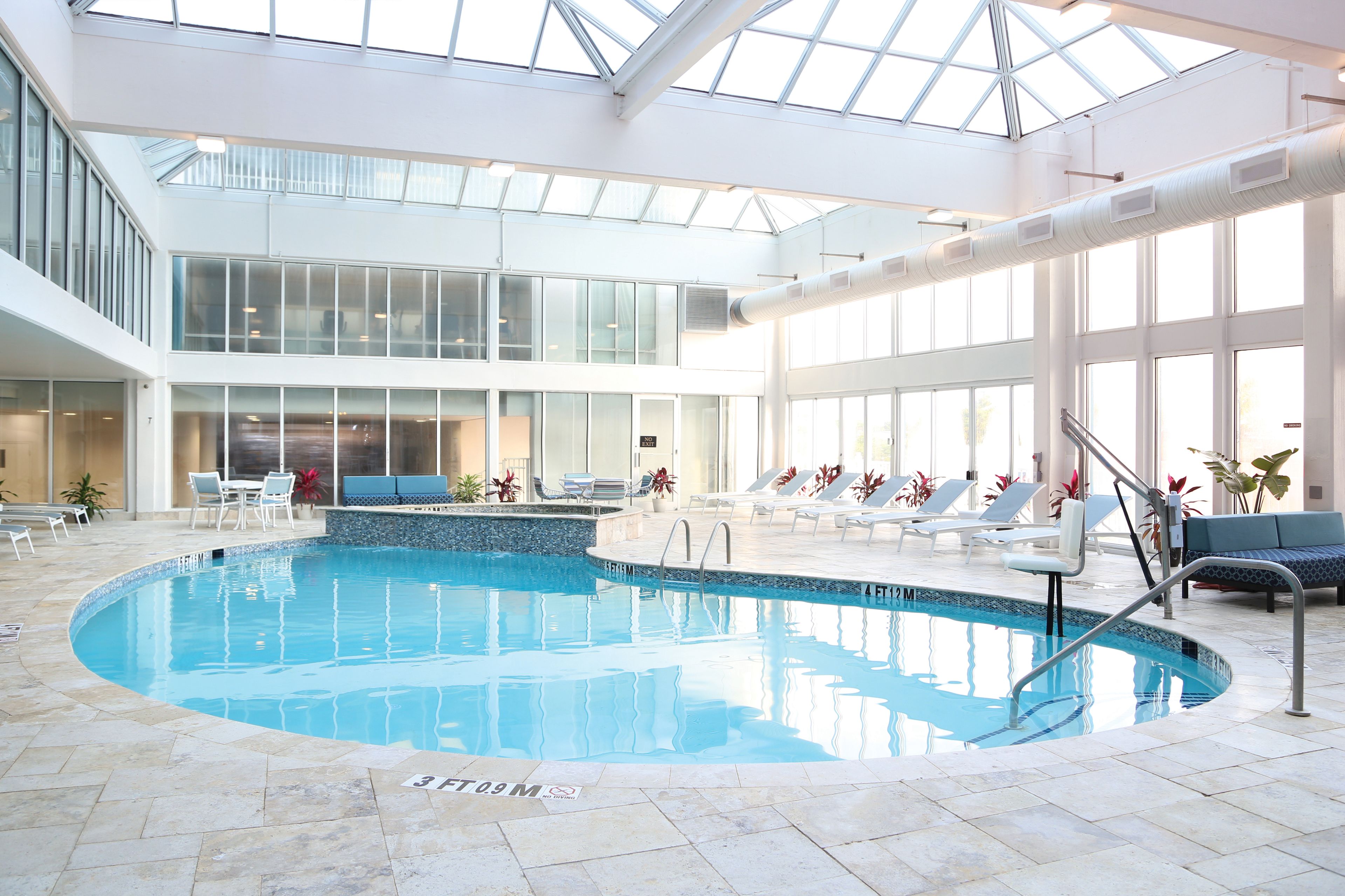 Una piscina interior con techos de vidrio transparente, sillas de descanso y un área de estar.