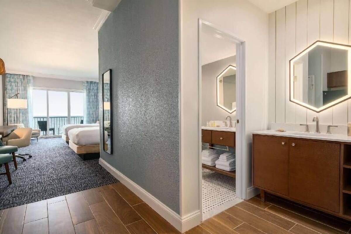 A hotel room with a bed, a desk, a chair, a bathroom with a mirror, and a view of the outdoors.